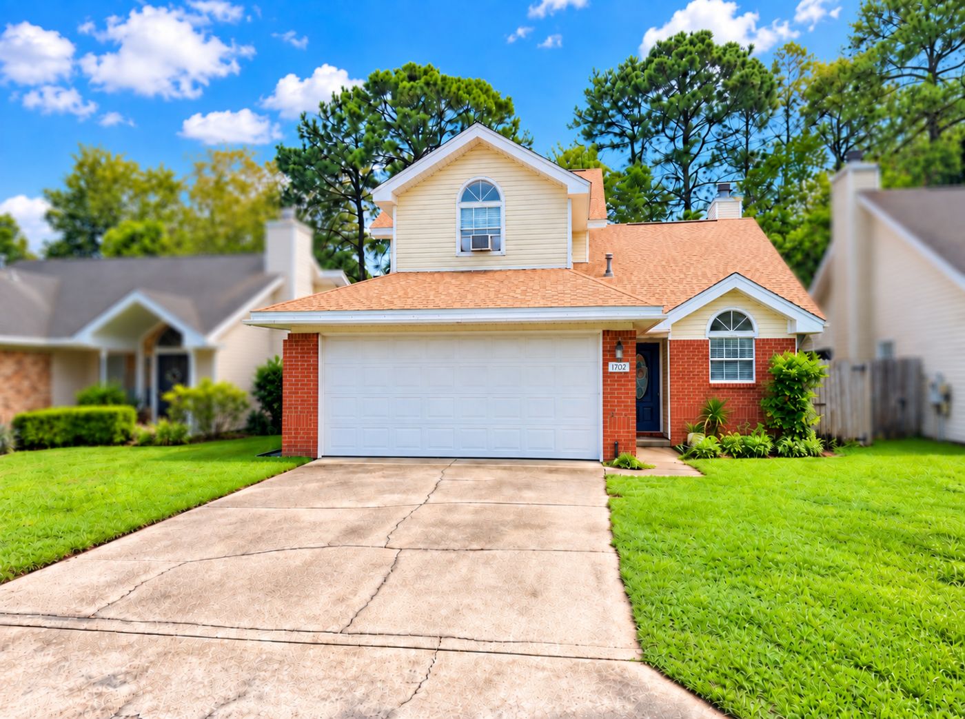 Front of 1702 Colonial Court: two-story home with red brick and cream siding, white two-car garage, navy front door and a green front lawn under tall pines